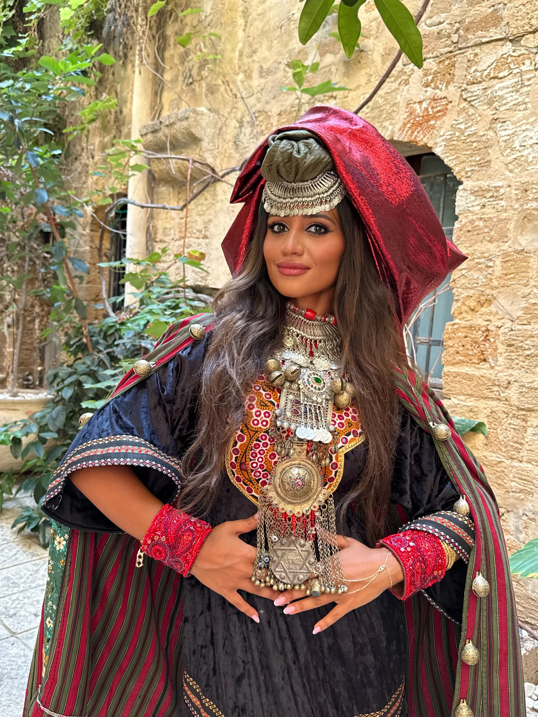 Yzabela standing in a stone courtyard in a black garment, striped mantle, red cuffs and layered necklaces